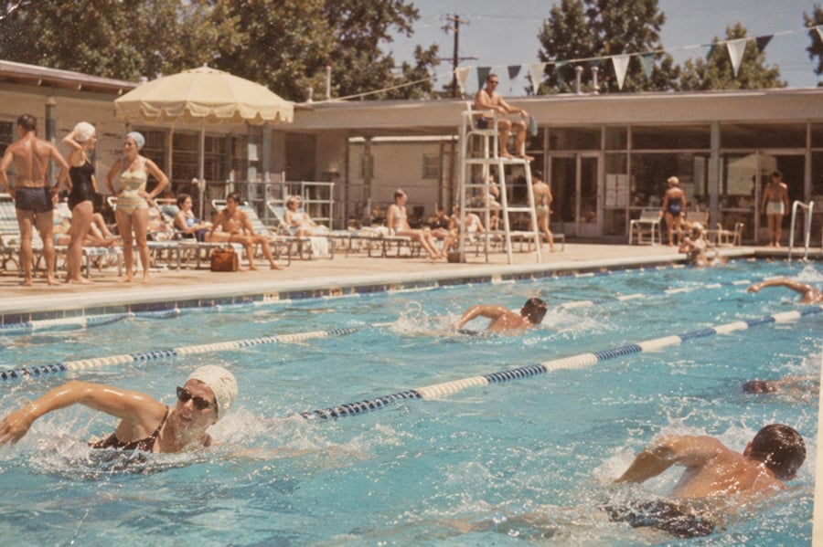 Swimmers in a pool race while people watch from the deck. A lifeguard sits on a high chair, and sunbathers relax under umbrellas. The setting appears to be a community pool on a sunny day.