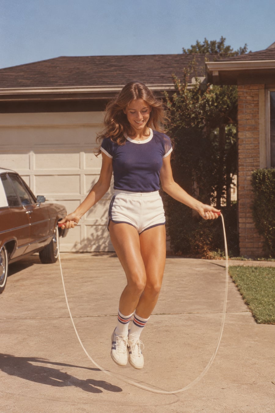 A woman in athletic clothing is jumping rope outside on a driveway, smiling. She wears a navy t-shirt, white shorts, tube socks, and sneakers. There’s a car and a garage in the background on a sunny day.