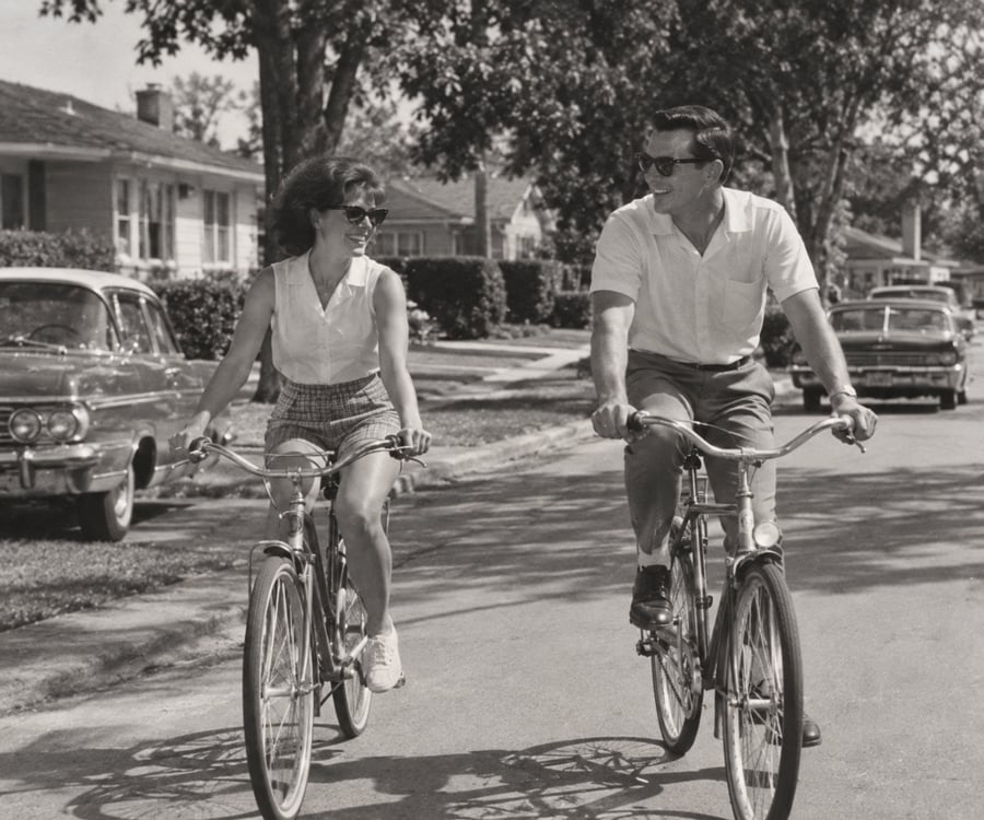 A man and woman ride bicycles side by side on a suburban street, smiling at each other. Vintage cars and houses line the street under leafy trees. The photo is black and white.