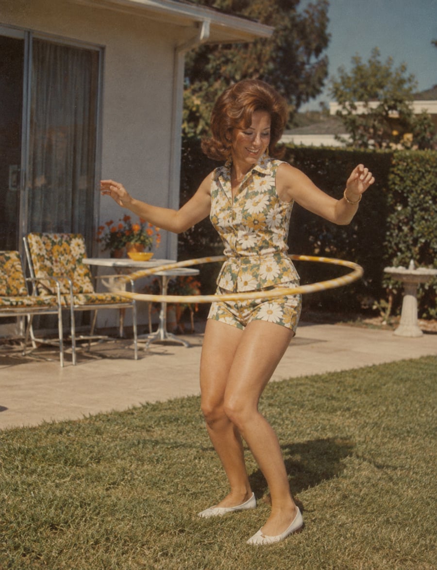 A woman in a floral outfit plays with a hula hoop on a grassy lawn, smiling. Patio furniture with floral cushions and a potted plant are visible near a house in the background.