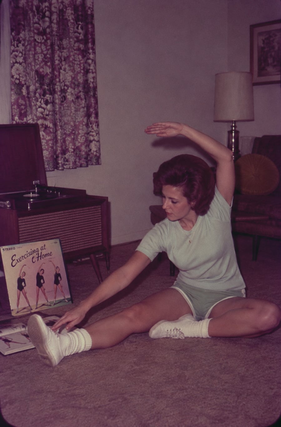 A woman in workout clothes stretches on a carpeted floor beside a record player and an “Exercising at Home” vinyl album in a vintage living room.