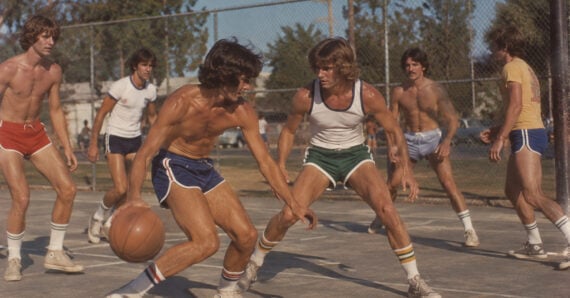 Six young men play basketball outdoors on a sunny day, wearing retro athletic shorts and tank tops. Trees and a chain-link fence surround the court.