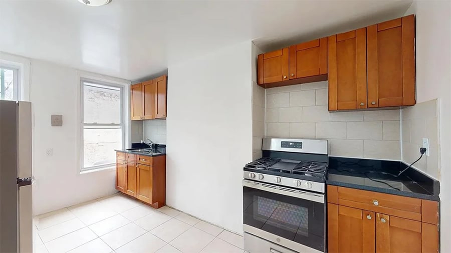 Bright kitchen with wood cabinets, black countertops, a stainless steel gas stove, and a window letting in natural light. Light-colored tile floor and walls give the room a clean, spacious feel.