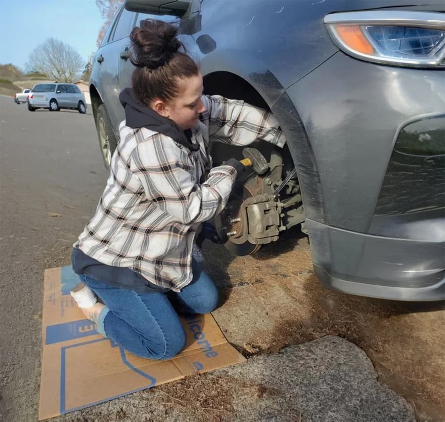 A person kneels on cardboard next to a car, using tools to work on the exposed wheel assembly. The car is parked on a paved area, and there are other cars and trees in the background.