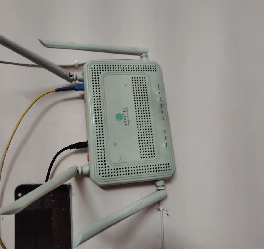 A white wireless router with three antennas is mounted on a wall, with multiple cables connected to it. Green indicator lights are visible on the front panel.