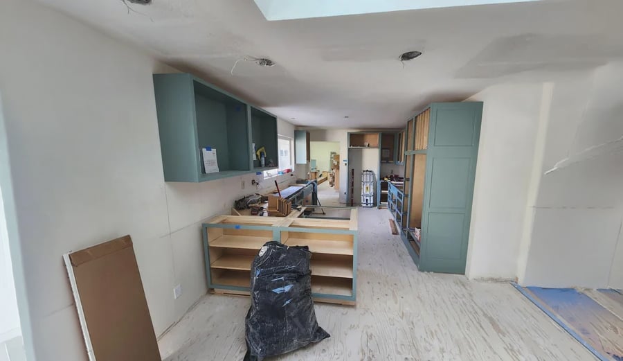 A kitchen under renovation with unfinished cabinets, exposed shelves, and tools scattered around. The floor is bare, and a black plastic bag is in the center of the room. Walls and ceiling are unpainted.