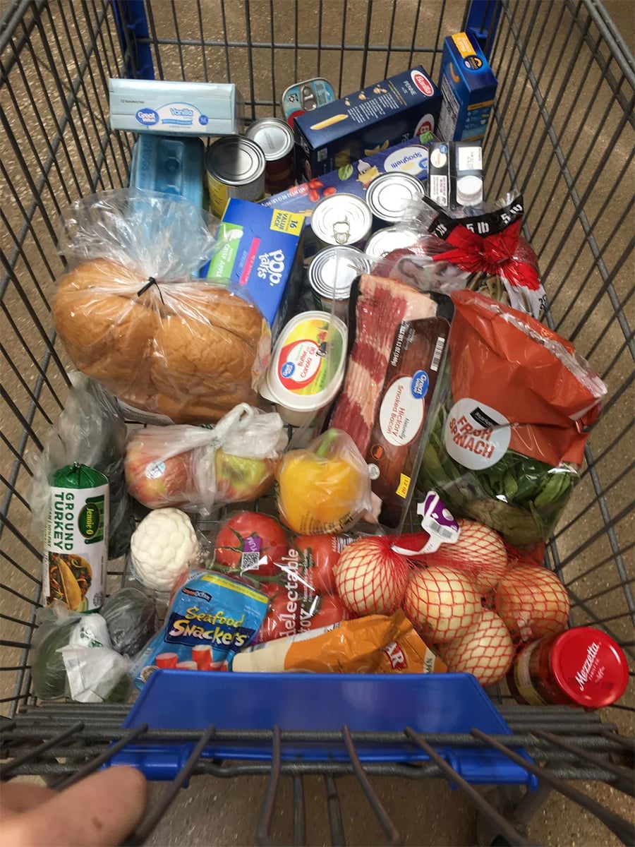 A shopping cart filled with groceries, including bread, canned goods, bacon, produce, cheese, eggs, packaged snacks, and various vegetables, viewed from above. A person’s hands are visible holding the cart handle.