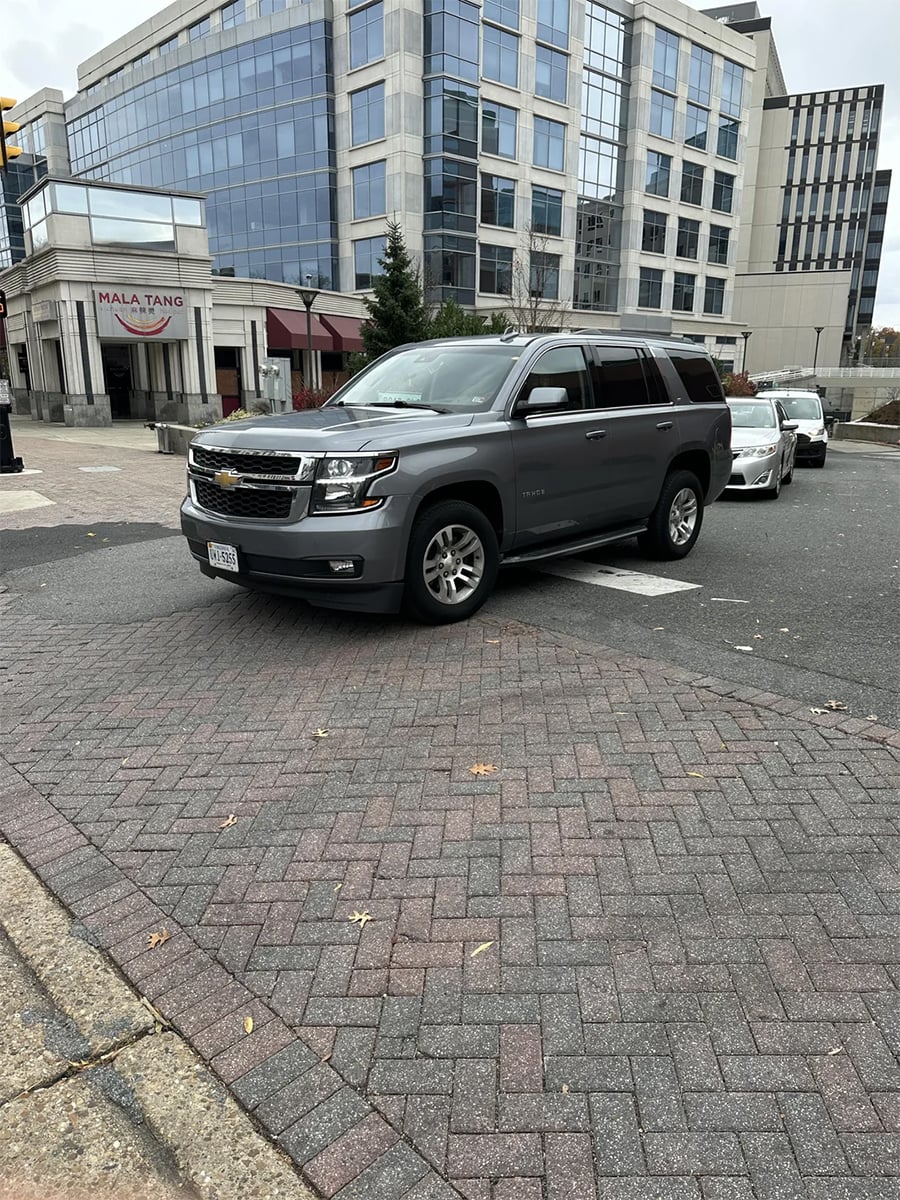 A gray SUV is parked on a brick-paved sidewalk near a busy street, in front of a modern glass office building and a restaurant called MALA TANG.