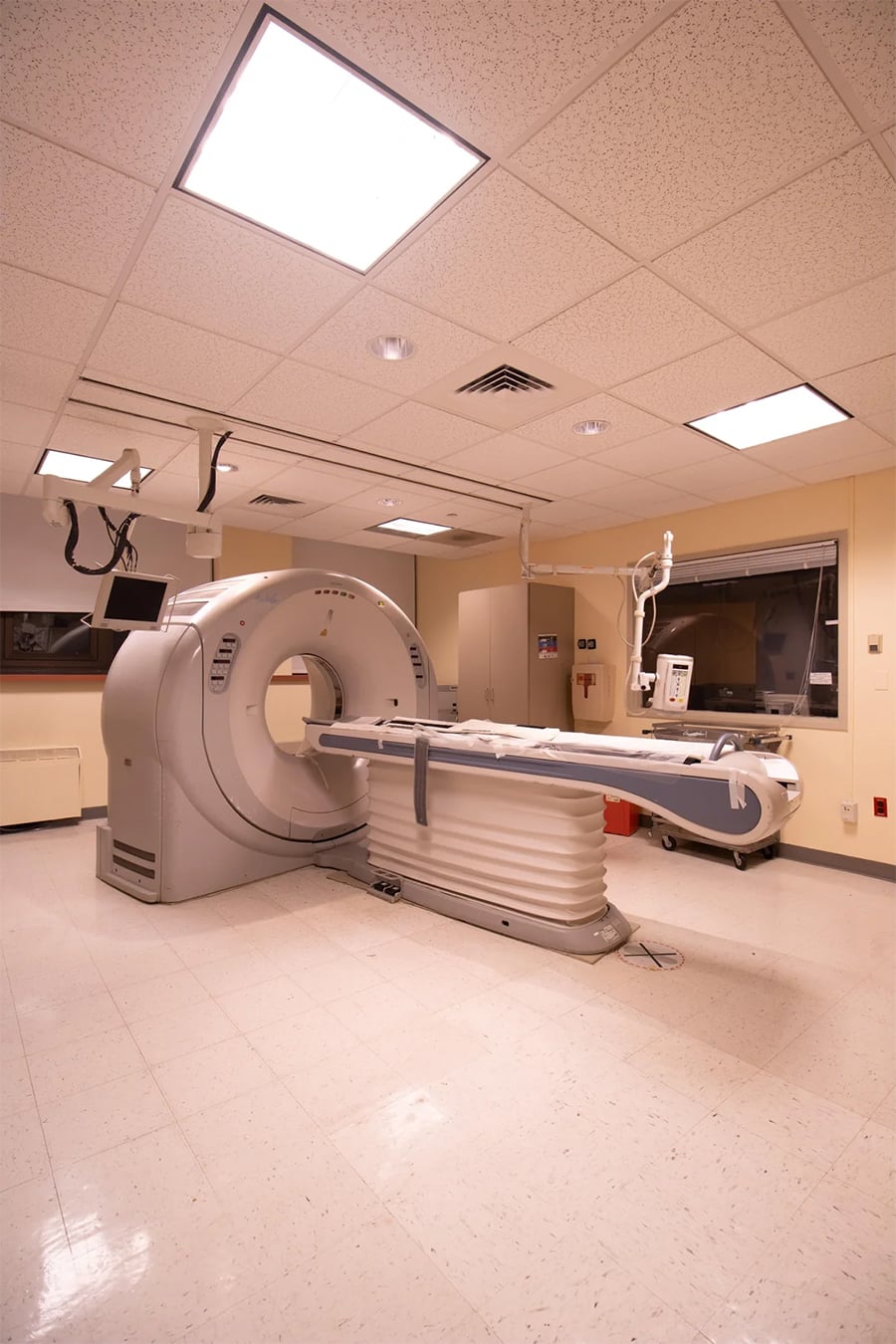 A CT scanner is positioned in the center of a hospital room with bright ceiling lights, beige walls, and medical equipment nearby.