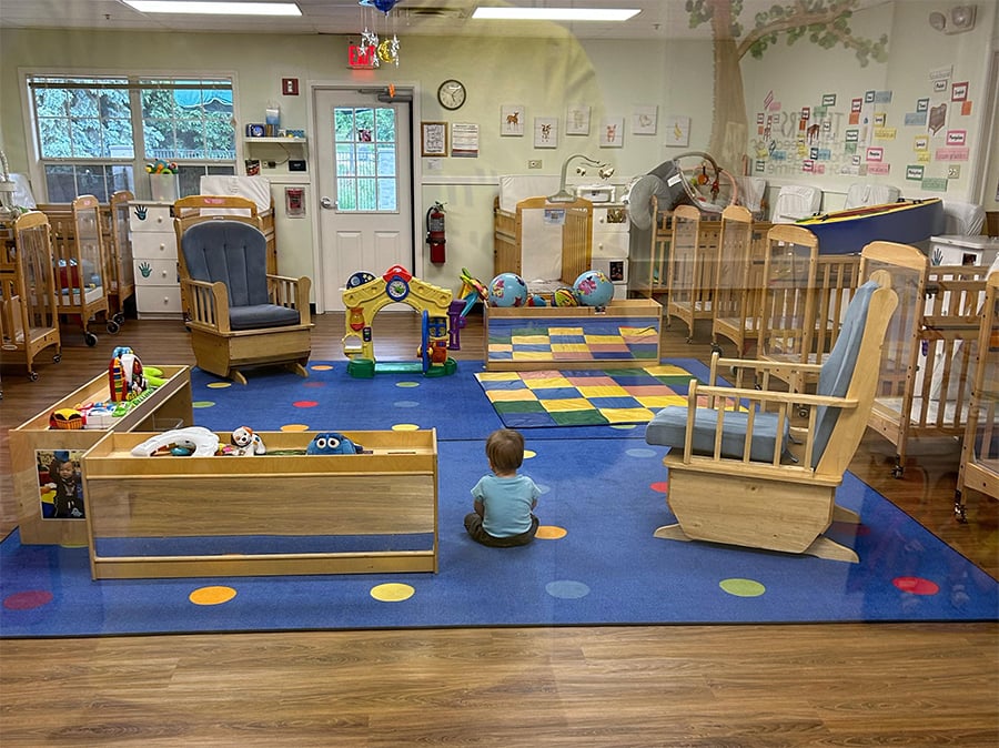 A toddler sits alone on a colorful rug in a daycare room filled with wooden cribs, toys, rocking chairs, and bright decorations.