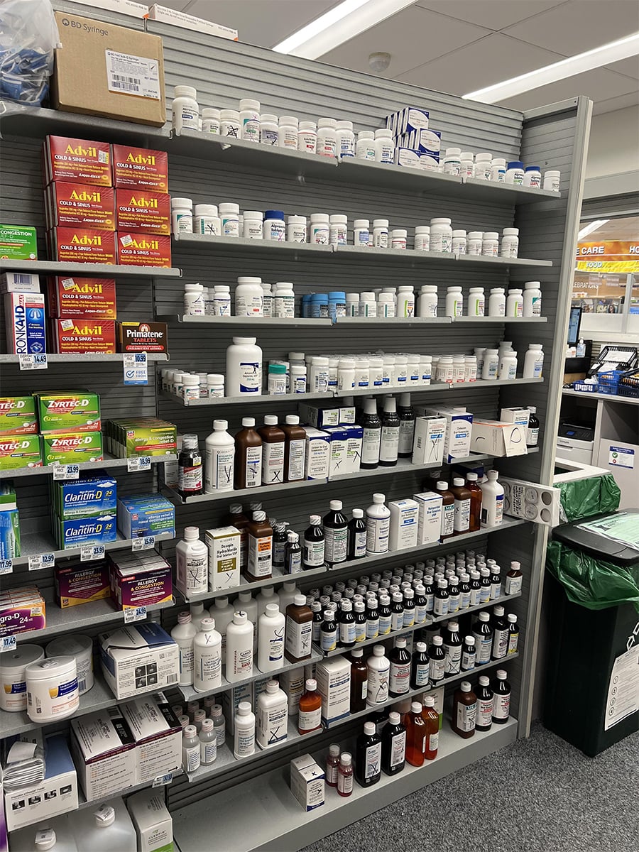 Shelves in a pharmacy stocked with various prescription medication bottles, pill containers, and over-the-counter medicine boxes, including Advil. The shelves are neatly organized, and the pharmacy counter is visible in the background.