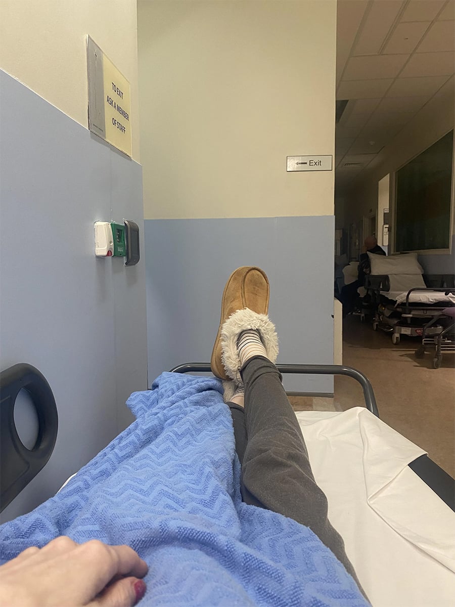 A person lies on a hospital bed covered with a blue blanket, wearing gray pants and tan slippers. Hospital hallway and exit sign are visible in the background.