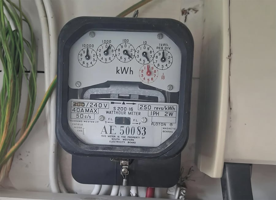 A close-up of an analog electricity meter mounted on a wall, displaying several dials and measurement labels, with electrical wires visible to the left side.