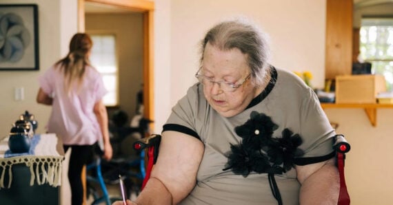 An elderly woman sits in a wheelchair, writing or drawing on paper, while a caregiver walks in the background inside a bright, cozy home.