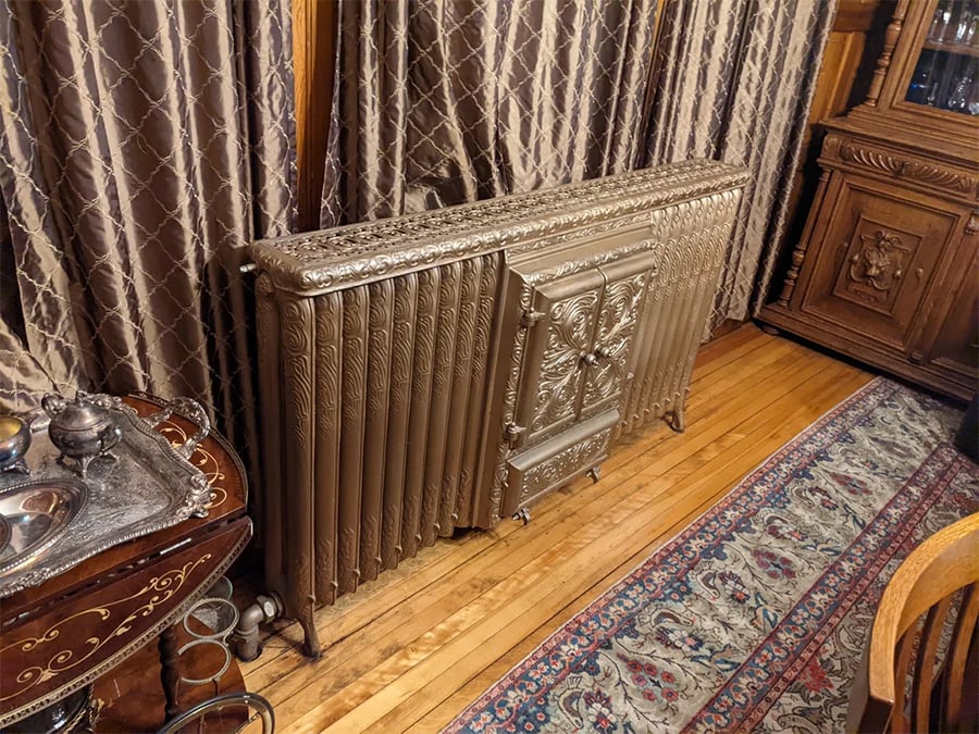 An ornate, vintage metal radiator with decorative detailing stands on a hardwood floor beside patterned curtains, a wooden cabinet, and a traditional rug.