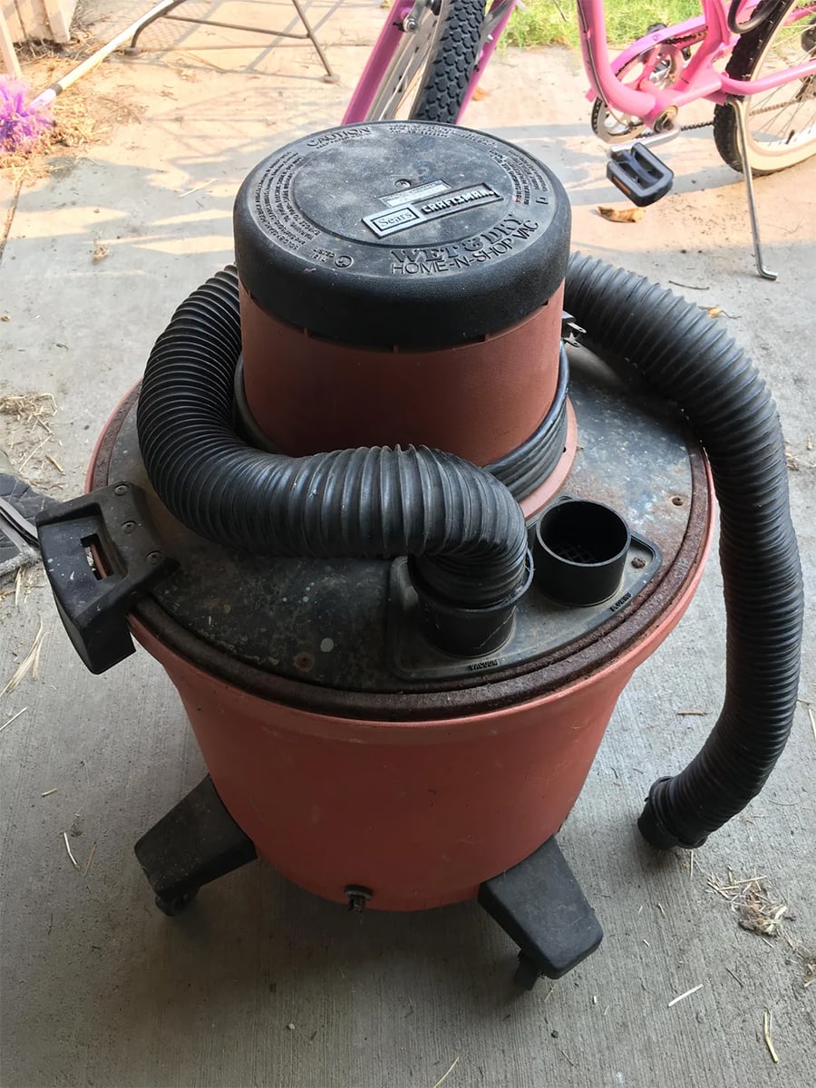 A red and black wet/dry shop vacuum sits on a concrete surface with a bike in the background. The vacuum has a coiled hose and wheels at its base.