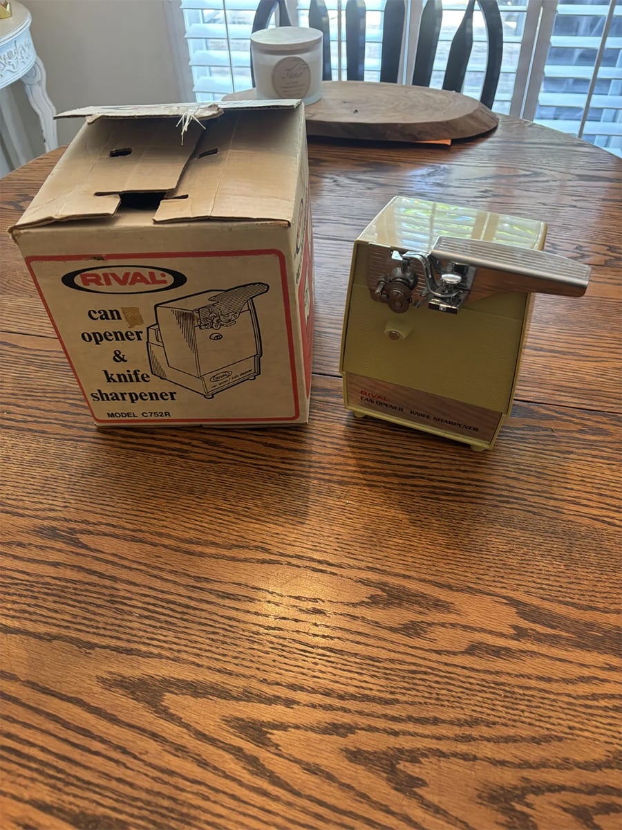 A Rival can opener and knife sharpener sits next to its box on a wooden table. The can opener is yellow with a silver lever, and the box shows an illustration and product details.