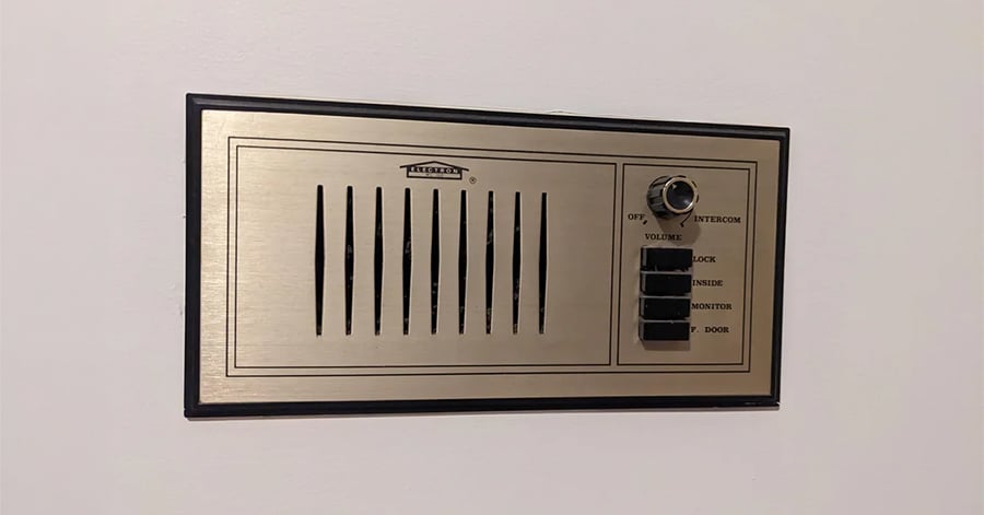 A vintage wall-mounted intercom panel with a speaker grille, volume knob, and labeled buttons for various functions, set against a plain white wall.