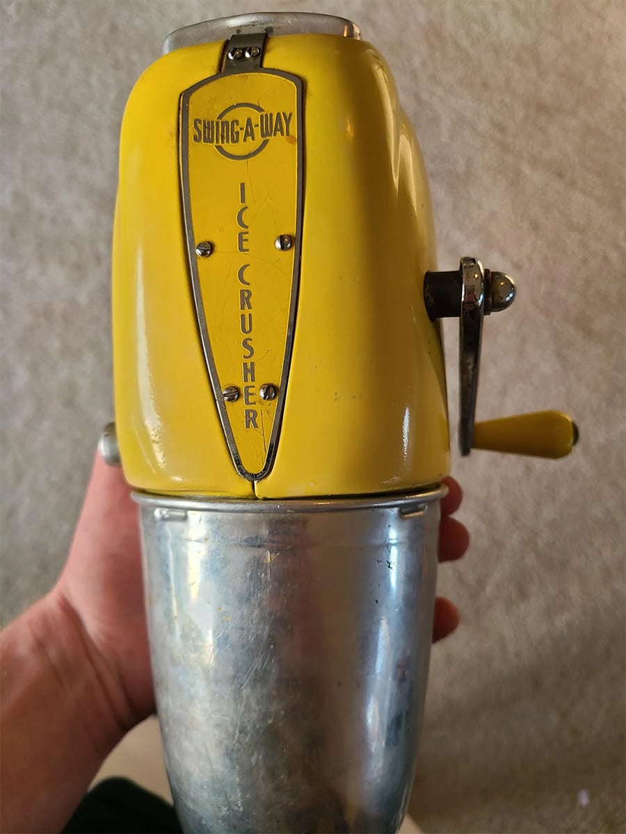 A vintage yellow Swing-A-Way ice crusher with a metal crank handle and silver bottom, held in someone’s hand against a neutral background.