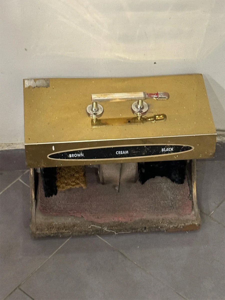 A vintage shoe polishing machine with labeled sections for brown, cream, and black polish stands on a tiled floor. Brushes and a gold-colored top are visible.