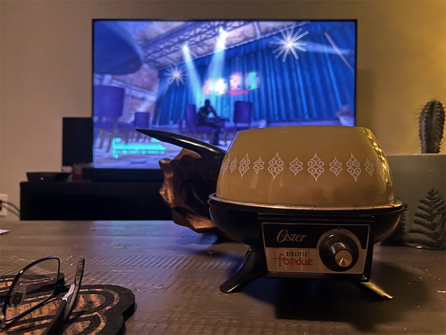 A vintage Oster electric fondue pot sits on a table in front of a TV displaying a video game scene. Glasses and a plant are nearby.