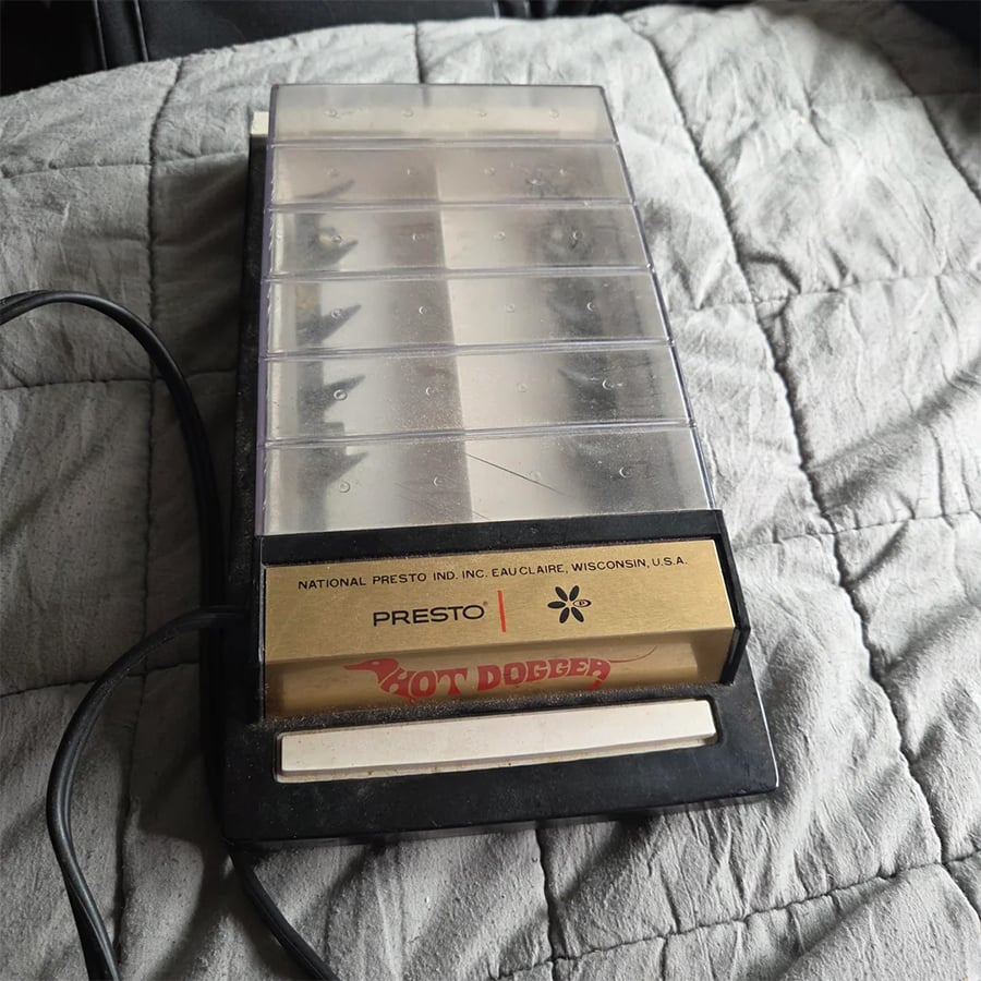 A vintage Presto Hot Dogger appliance sits on a gray quilted bedspread, with a power cord visible to the side. The top cover is transparent, showing slots underneath.