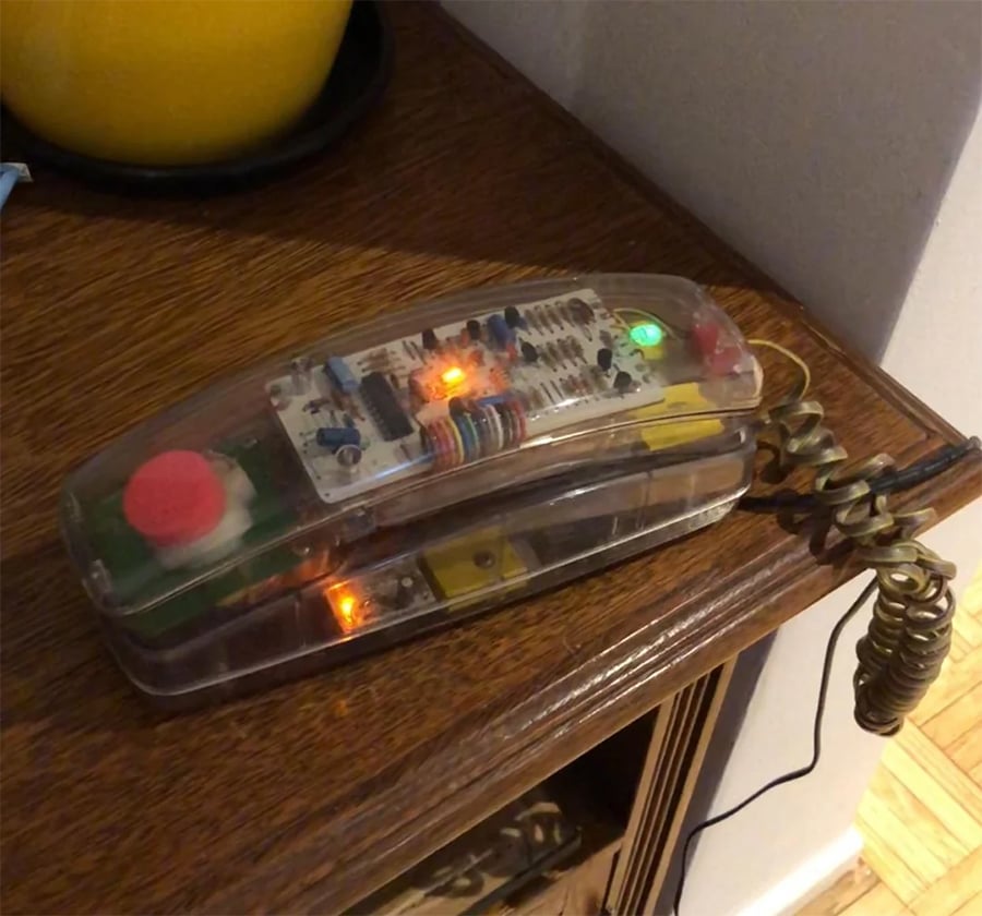 A transparent landline phone with visible internal electronics sits on a wooden table, illuminated by green and orange lights with a coiled cord attached.