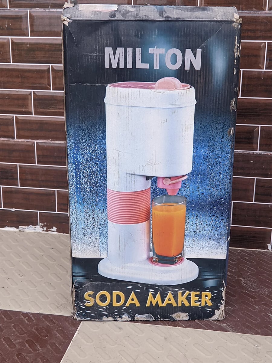 A box with “MILTON SODA MAKER” printed on it shows an image of a white and pink soda maker dispensing orange drink into a glass, against a blue gradient background.