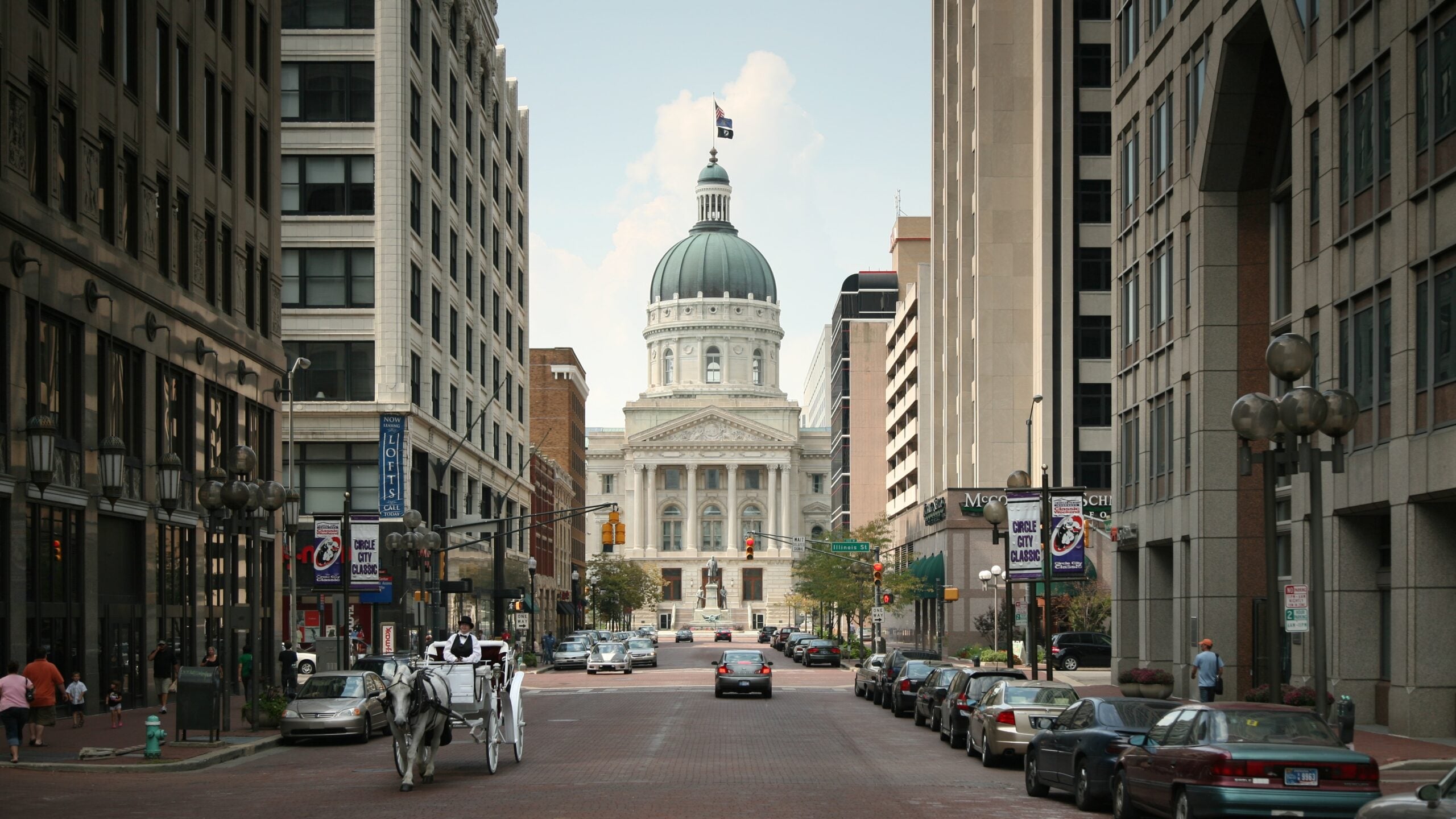 Street view of a domed government building flanked by tall offices, with cars parked along the road and a horse-drawn carriage traveling down the brick-paved street toward the building.
