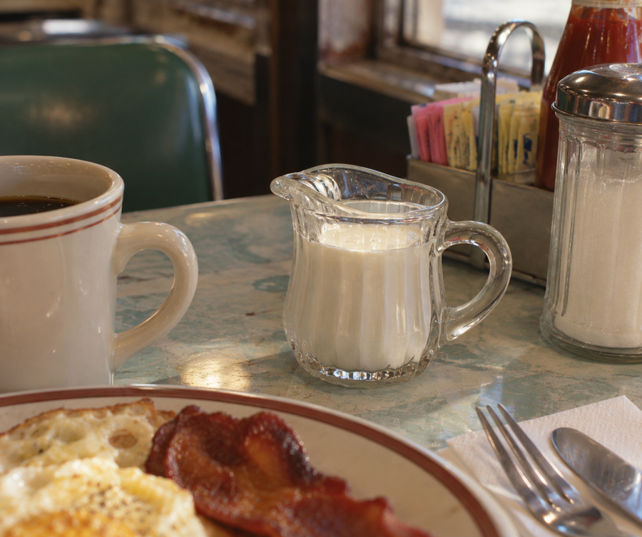 Glass jug of cream, coffee cup, plate with eggs and bacon, napkin with cutlery, and containers of sugar and sweetener packets on a diner table.
