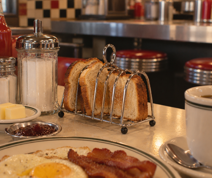 Slices of toast in a metal rack, a sugar jar, and a plate with fried eggs and bacon on a diner counter, with red stools and a checkered wall in the background.