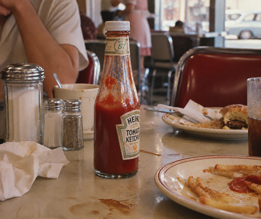 A Heinz ketchup bottle sits on a diner table with fries, a half-eaten burger, salt and pepper shakers, and a napkin. A person’s arm is visible, and red chairs and other diners are in the background.