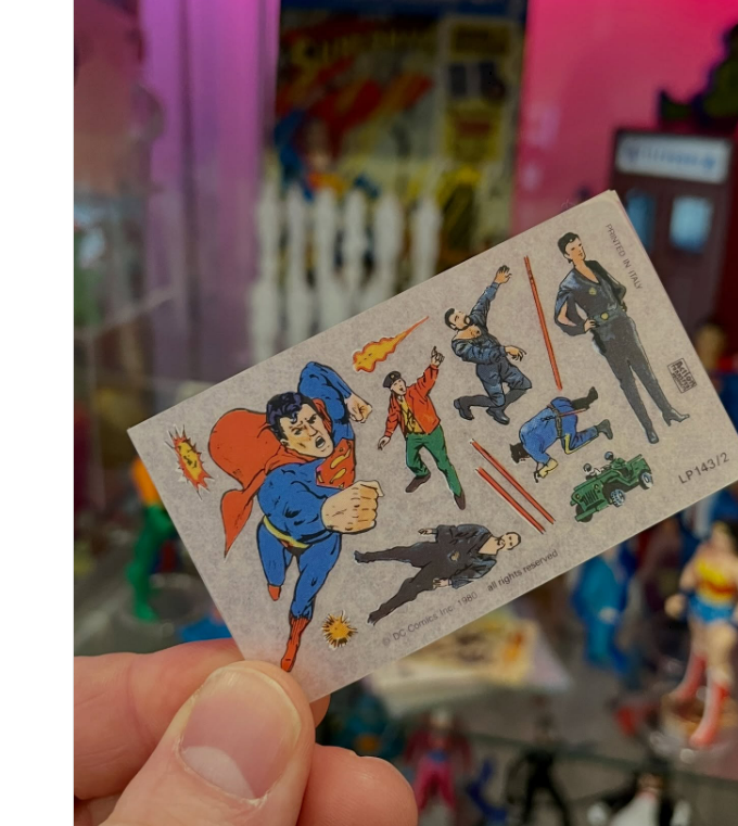 A hand holds a small card featuring vintage-style illustrations of Superman in various action poses, with a colorful display of comic book memorabilia blurred in the background.