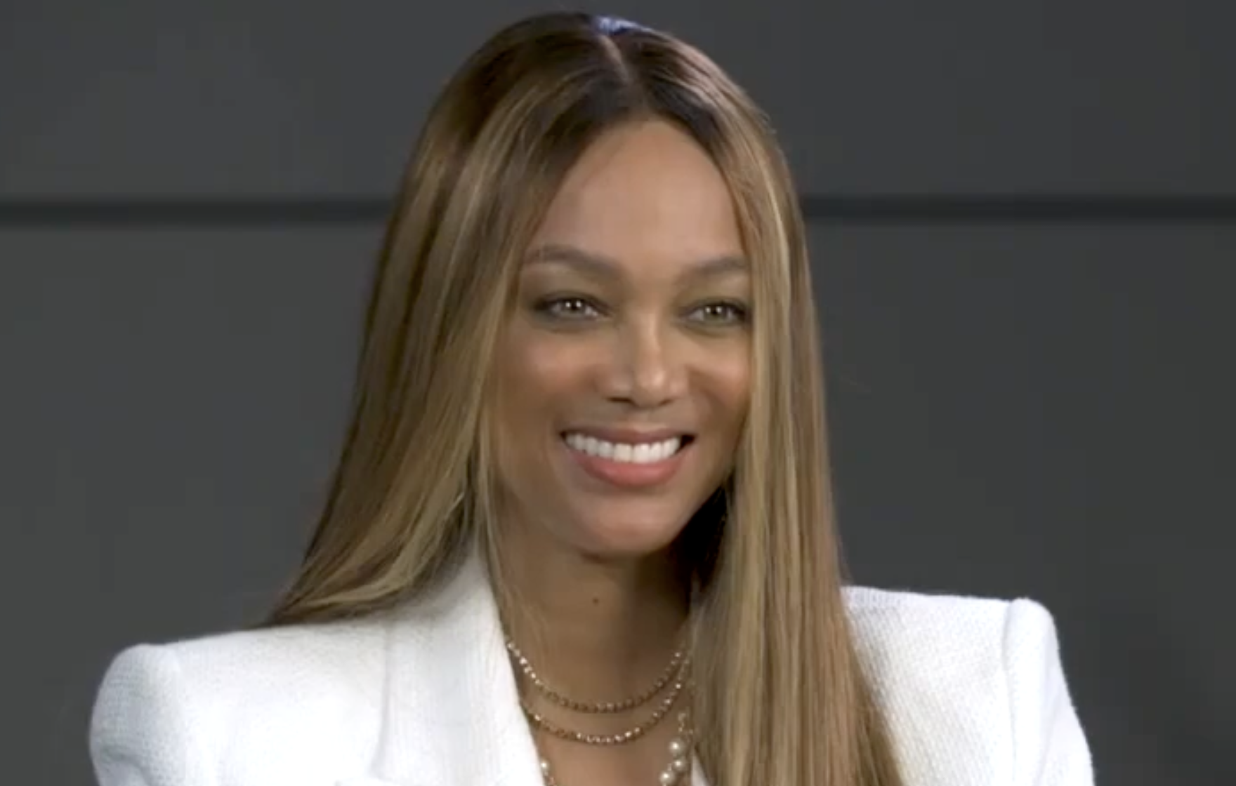 A woman with long straight hair and a white blazer smiles in front of a dark background.