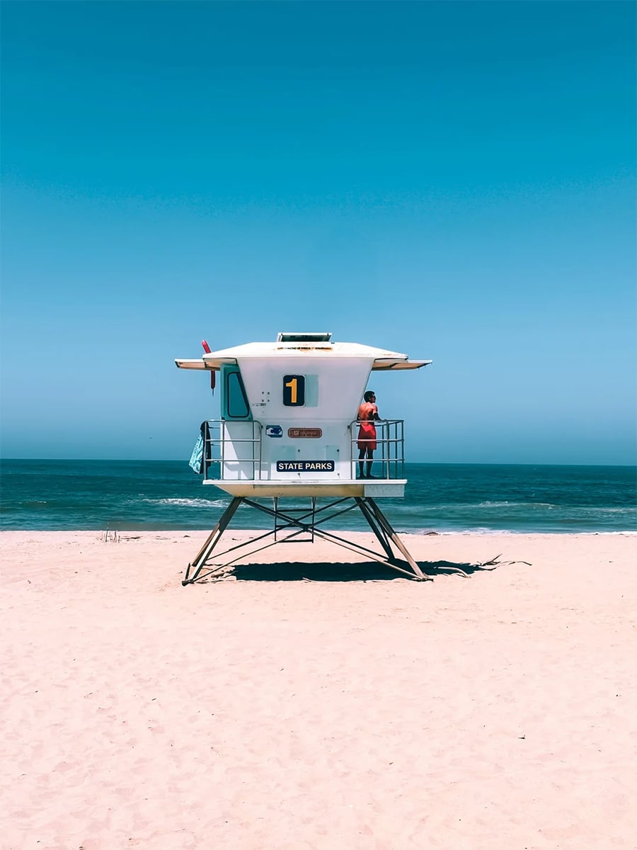 A lifeguard tower with the number 1 sits on a sandy beach by the ocean. A person in red stands on the balcony, looking at the water under a clear blue sky.