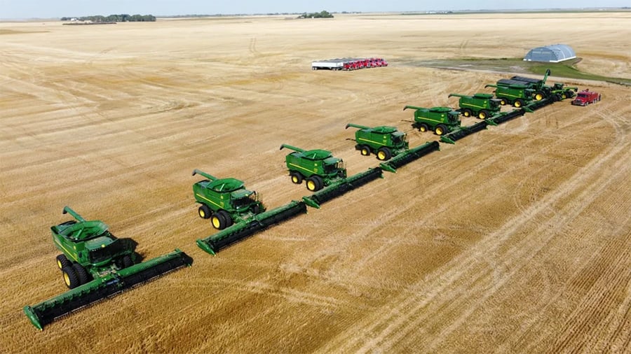 Seven green combine harvesters are lined up diagonally across a large, harvested wheat field, with equipment, vehicles, and a metal building visible in the distance under a clear sky.