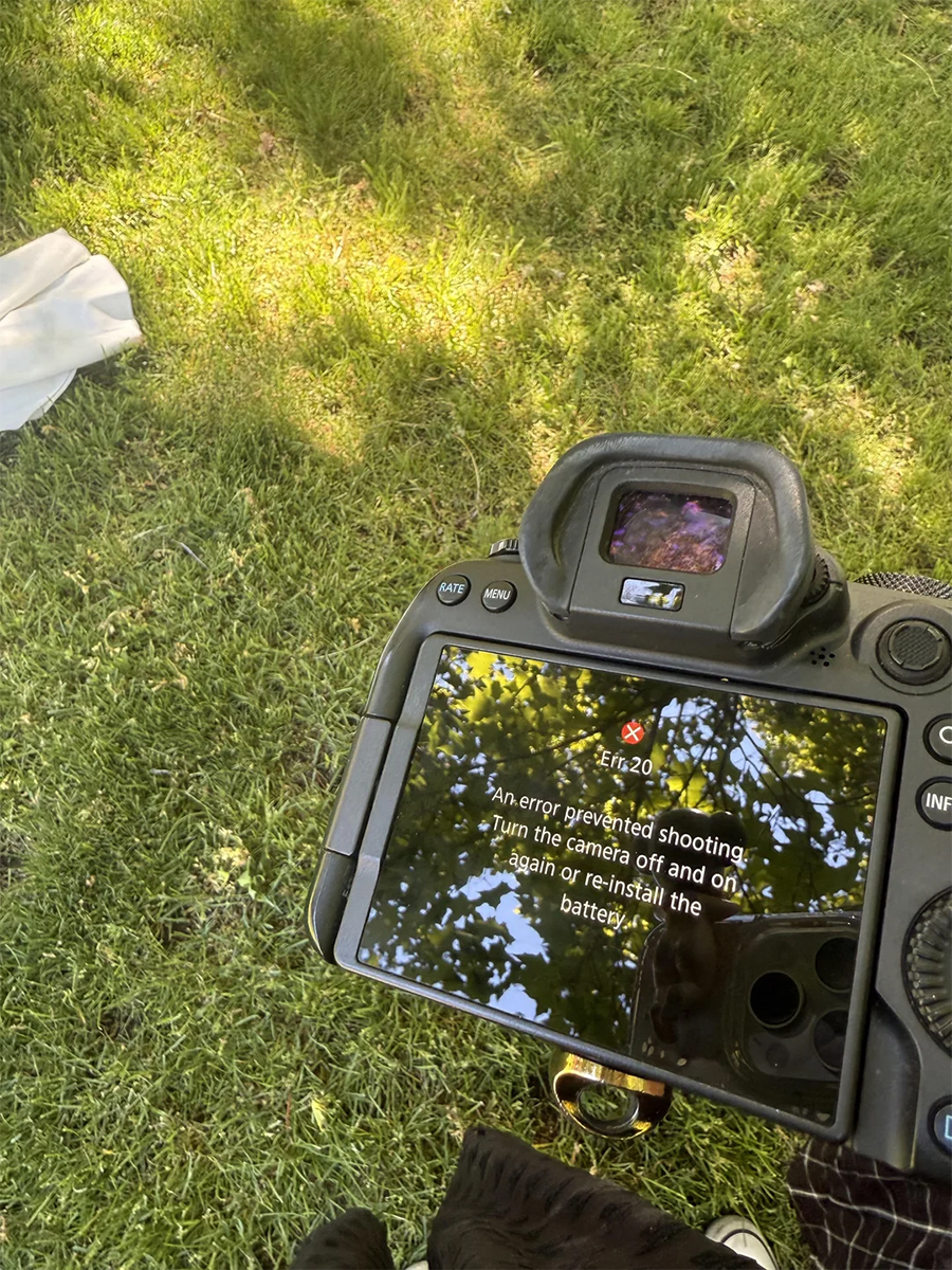 A camera screen displays an error message under a tree, with sunlight and leaves reflecting on it. Grass and part of a white cloth are visible on the ground.