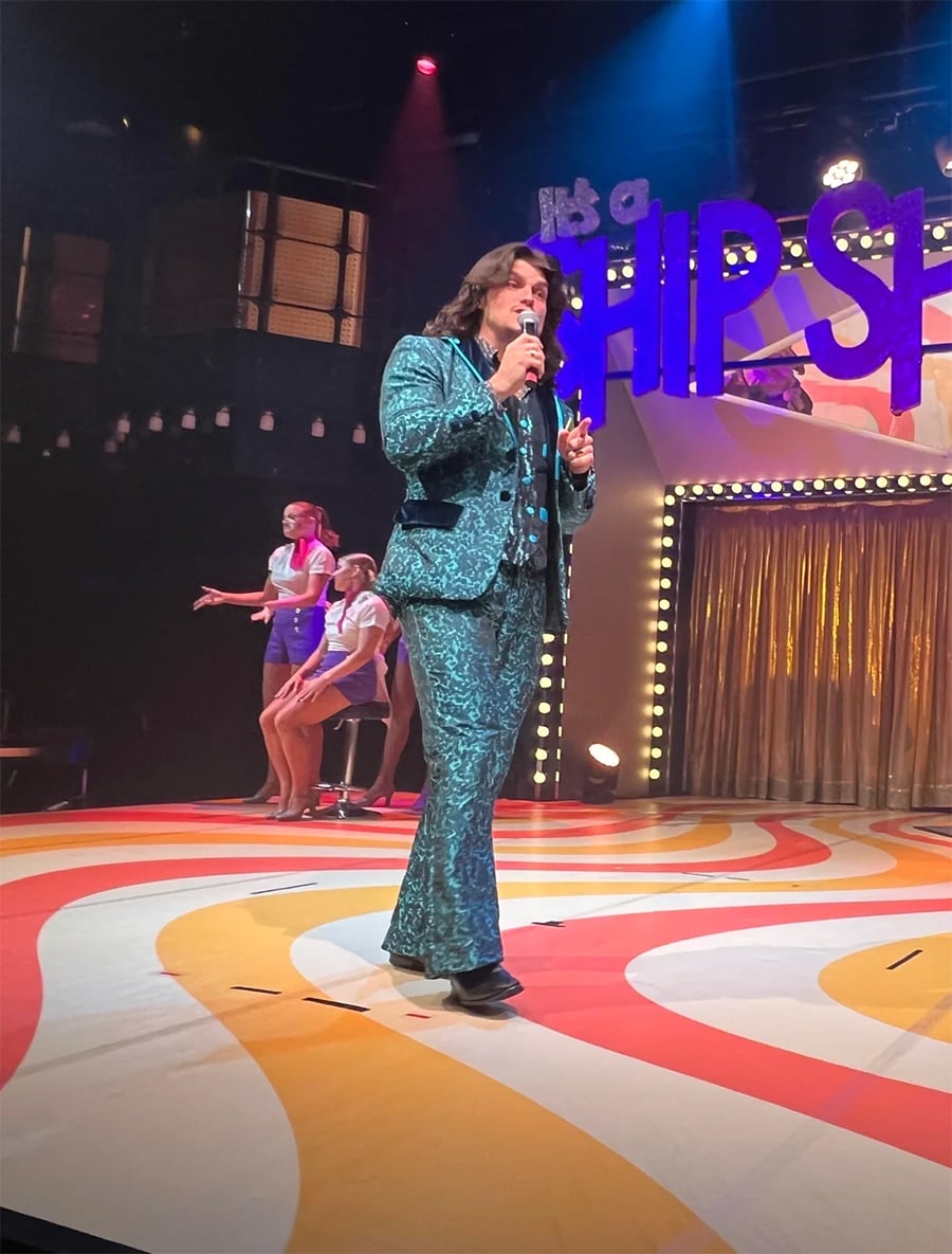 A man in a shiny teal suit sings on a colorful stage with “HIP” in large letters behind him. Two women in retro outfits dance in the background under stage lights.