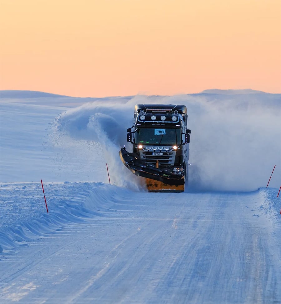 A snowplow truck clears a snow-covered road at sunset, with snow spraying from the plow as it works. The landscape is snowy and flat, and the sky is a soft orange and blue gradient.