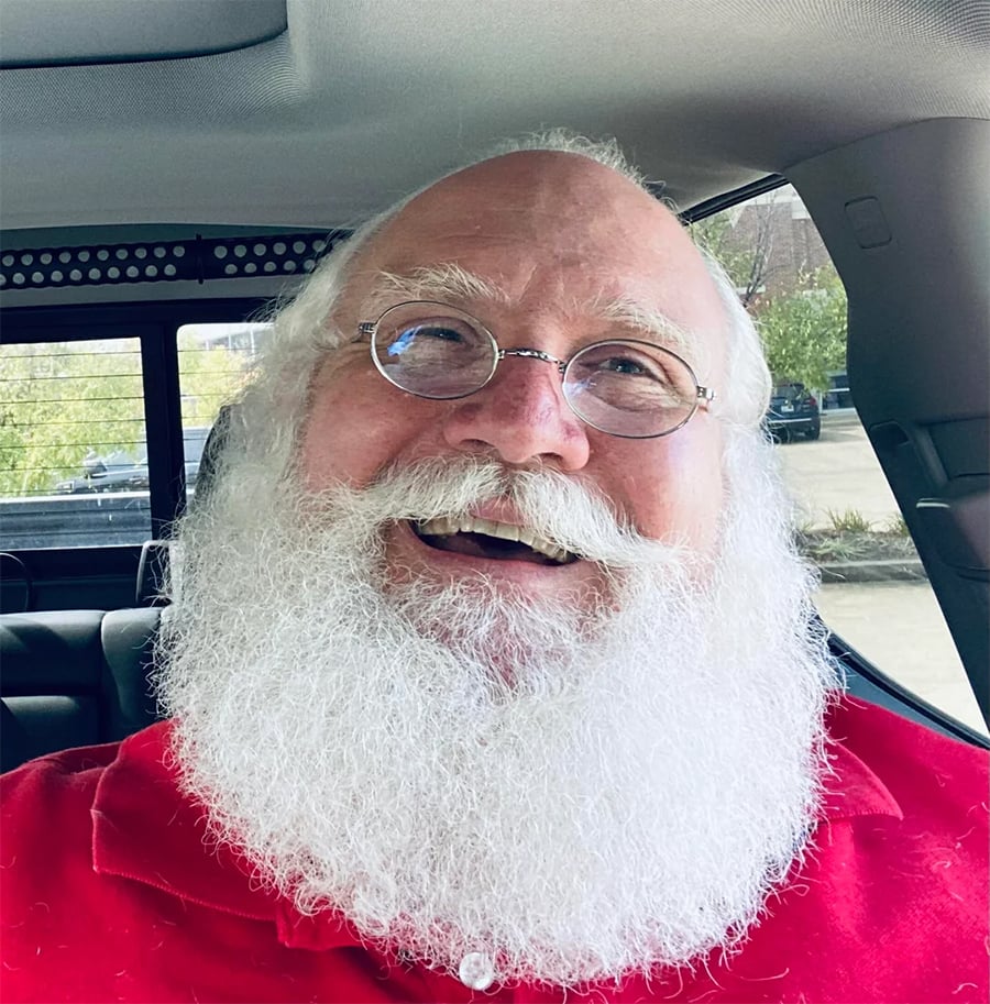 An older man with white hair, a full white beard, and round glasses smiles while sitting in a car, wearing a red shirt.