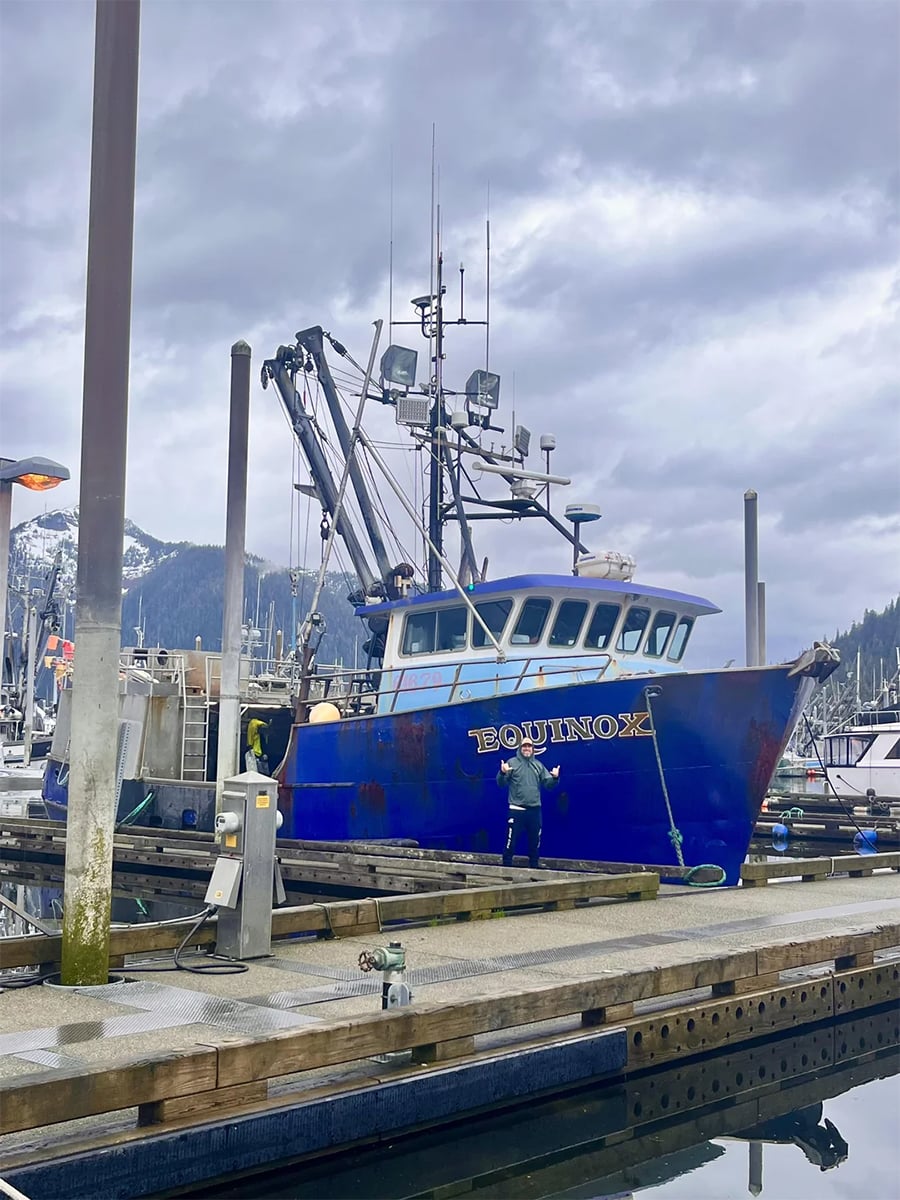 A blue fishing boat named “Equinox” is docked at a marina on a cloudy day. A person stands near the boat, and snowy mountains are visible in the background.
