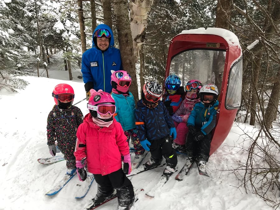 A group of young children in colorful ski gear and an adult stand in the snow beside a small red ski gondola, surrounded by trees in a snowy forest.