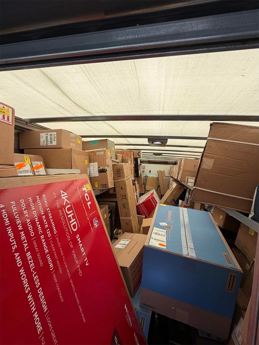 A delivery truck interior packed tightly with large boxes, including a visible TCL 4K UHD TV box and other assorted packages stacked up to the roof.