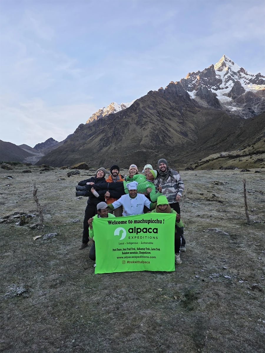 A group of people poses outdoors in front of snow-capped mountains, holding a green “Alpaca Expeditions” banner that says “Welcome to Machu Picchu!” The scene is at sunrise or sunset in a rugged landscape.