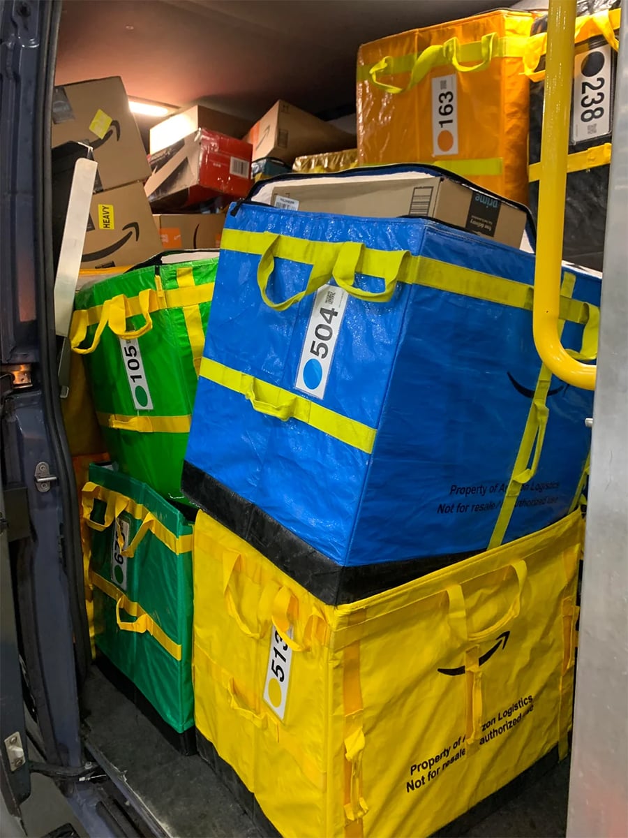 Colorful Amazon delivery bags and boxes are stacked and packed tightly inside a vehicle, labeled with various numbers and barcodes for sorting and identification.