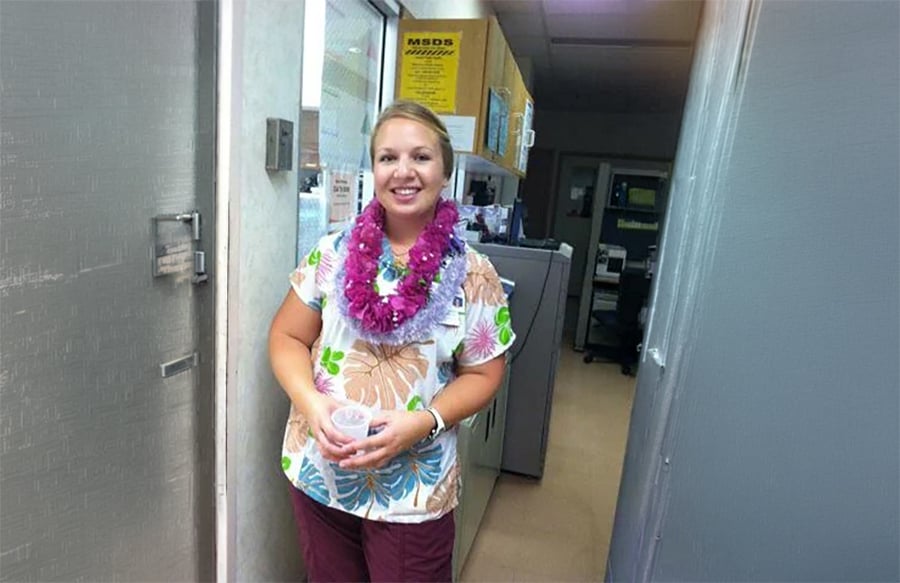 A woman in floral scrubs with a purple lei stands in a hallway, smiling and holding a small cup. She is near a door with an office area and cabinets visible in the background.