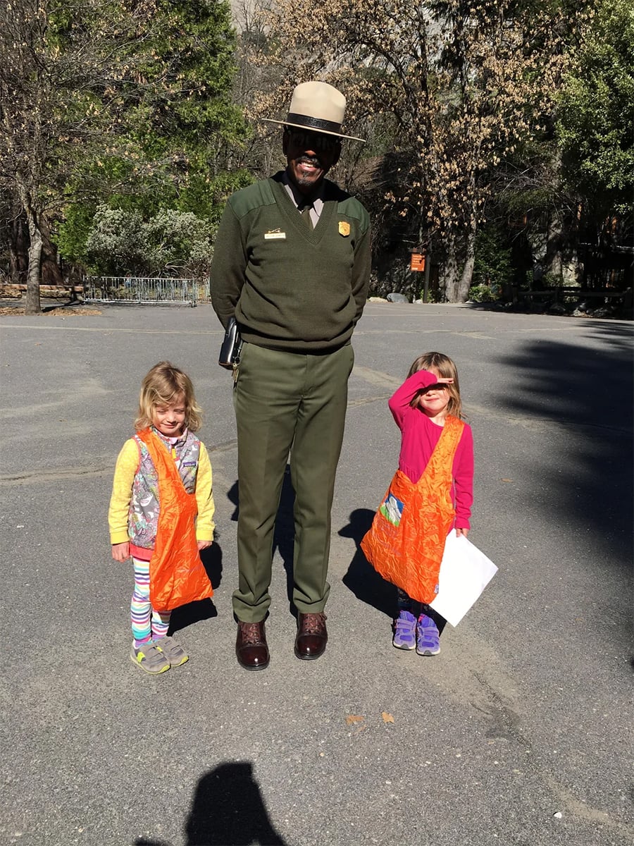 A park ranger stands smiling between two young children holding orange bags, one child shielding her eyes from the sun. They are outdoors with trees and pavement visible.