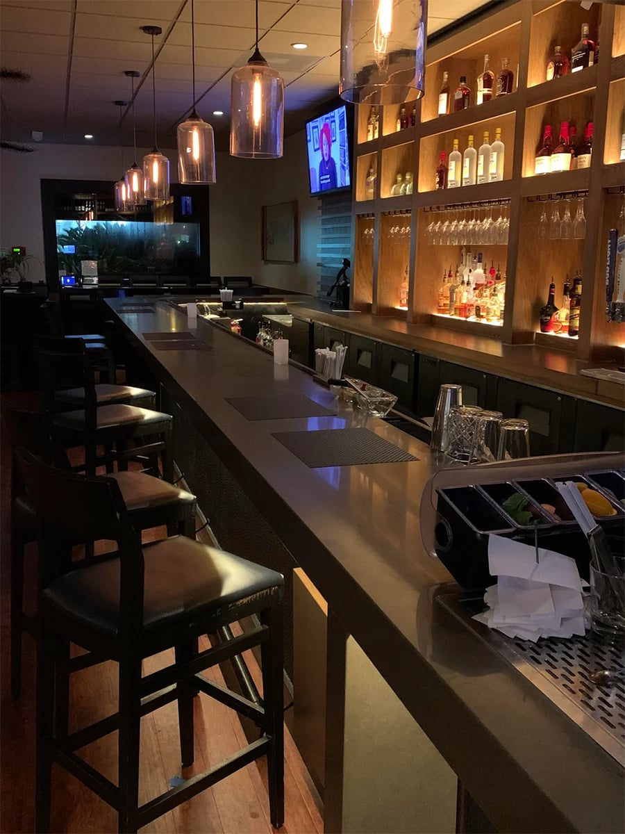 A modern bar with high chairs, hanging pendant lights, a stocked backlit shelf of bottles, and a TV on the wall. The bar counter has drink supplies and glasses.