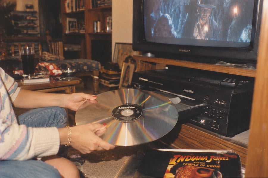 A person loads a large LaserDisc into a player. An Indiana Jones movie plays on the TV. An Indiana Jones: Temple of Doom LaserDisc case lies nearby on the carpet.