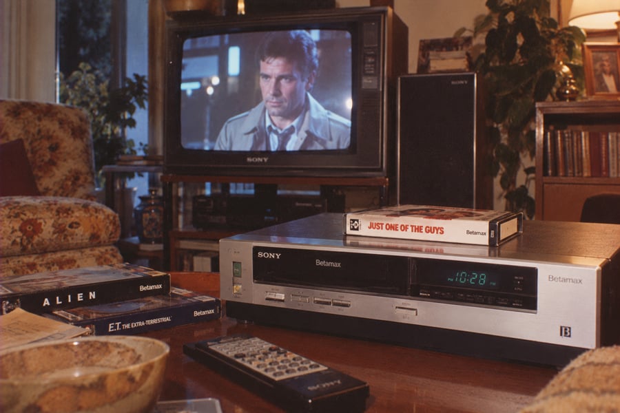 A living room scene with a Sony Betamax player, remote, Betamax tapes, and a TV showing a movie. Titles include “ALIEN,” “E.T.,” and “Just One of the Guys.” Cozy furniture and plants are visible in the background.